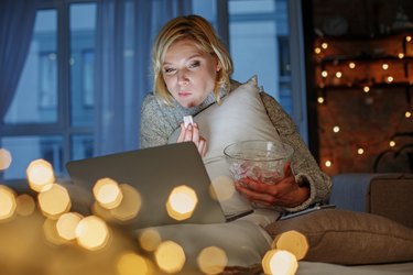 an adult with short blond hair holds a glass bowl of a healthy late night snack while watching a show on their laptop with string lights in the background