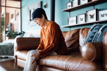 A senior Asian woman sitting on her couch at home and holding her leg because her legs feel weighted