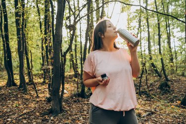 a person with shoulder-length brown hair wearing a pink t-shirt and drinking from a metal water bottle while walking in the woods