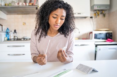 A person is holding a nasal swab and an at-home self-test cartridge in the kitchen at home after testing positive for covid-19